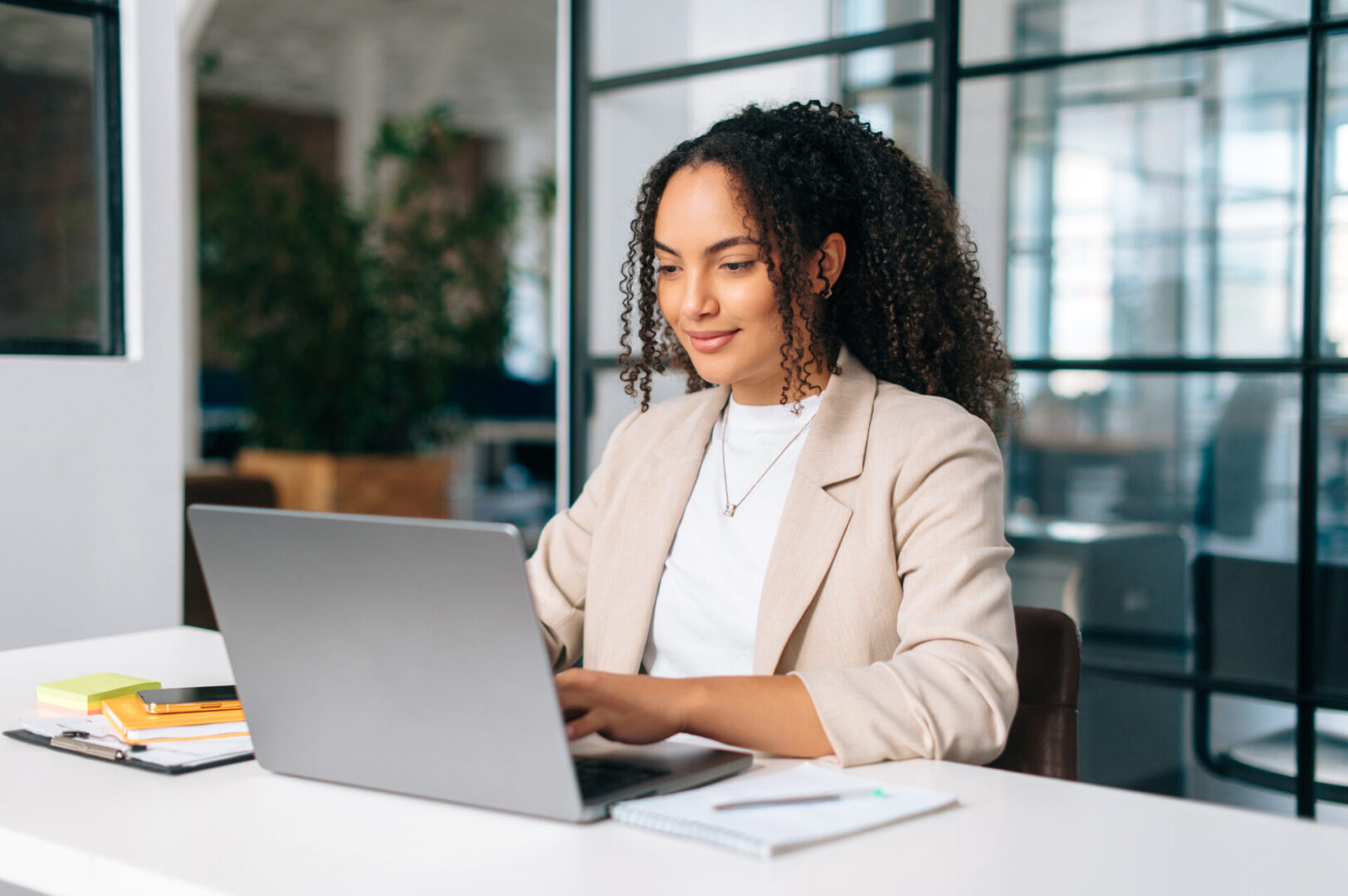 Woman uses laptop. Successful curly haired hispanic or brazilian woman in stylish elegant clothes, office employee, secretary or hr manager, using a laptop while sitting at her workplace, working Woman working on laptop in modern office.